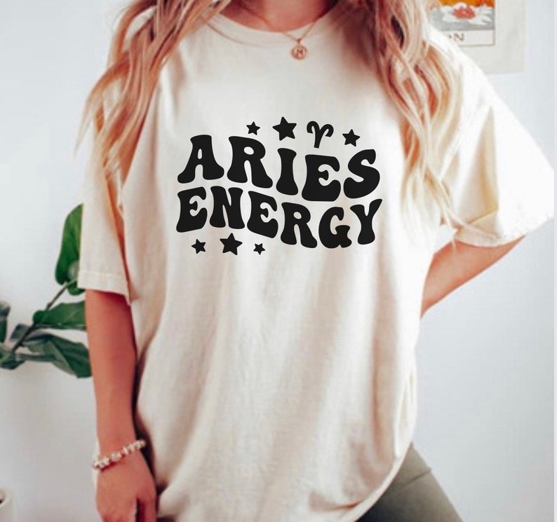 May include: Cream-colored t-shirt with the words "ARIES ENERGY" in bold, black, retro-style font. The design includes star and ram symbols. The shirt is a casual, oversized fit.