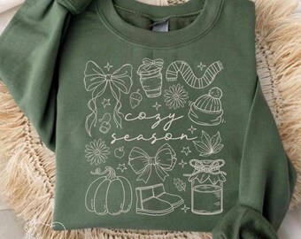 Könnte beinhalten: Grünes Sweatshirt mit weißen Strichzeichnungen von Schleifen, einer Kaffeetasse, einem Schal, einer Mütze, Blumen, einem Kürbis, Stiefeln und einem Glas. Die Worte "cozy season" sind kursiv geschrieben. Die Ärmel sind gerollt.