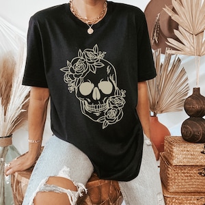 以下が含まれることがあります： 花柄の骸骨のグラフィックがプリントされた黒いTシャツ。骸骨はベージュで縁取られており、周りに花柄のデザインが施されています。
