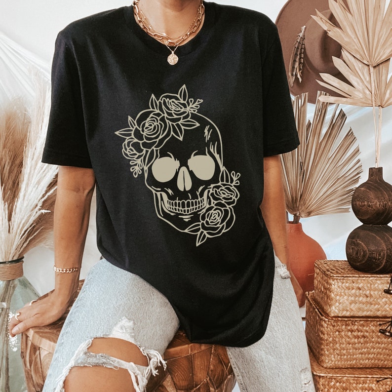 以下が含まれることがあります： 花柄の骸骨のグラフィックがプリントされた黒いTシャツ。骸骨はベージュで縁取られており、周りに花柄のデザインが施されています。