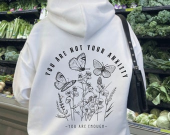 Può includere: Felpa bianca con una stampa grafica nera di farfalle e fiori. Il testo sulla felpa recita "You are not your anxiety - You are enough."
