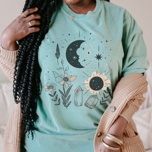 May include: A turquoise blue t-shirt with a graphic design of a crescent moon, sun, crystals, and flowers. The design is in black, white, and gold.