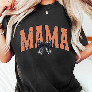 Puede incluir: Una camiseta negra con la palabra "MAMA" impresa en naranja con un lazo de encaje negro debajo.
