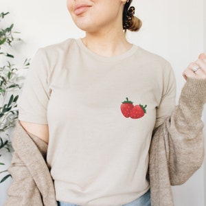Peut inclure: Un t-shirt beige avec un petit motif de deux fraises rouges sur la poitrine gauche.