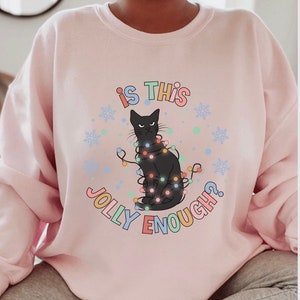 May include: A pink sweatshirt with a black cat wearing Christmas lights and the text "Is this jolly enough?" in colorful letters.
