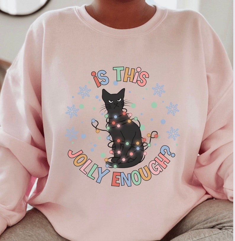 May include: A pink sweatshirt with a black cat wearing Christmas lights and the text "Is this jolly enough?" in colorful letters.