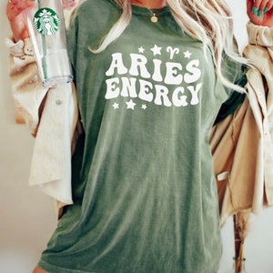 May include: Olive green t-shirt with the text "ARIES ENERGY" in white, featuring stars and the Aries symbol. The shirt has a vintage, slightly faded look. A clear Starbucks tumbler is held in the left hand.
