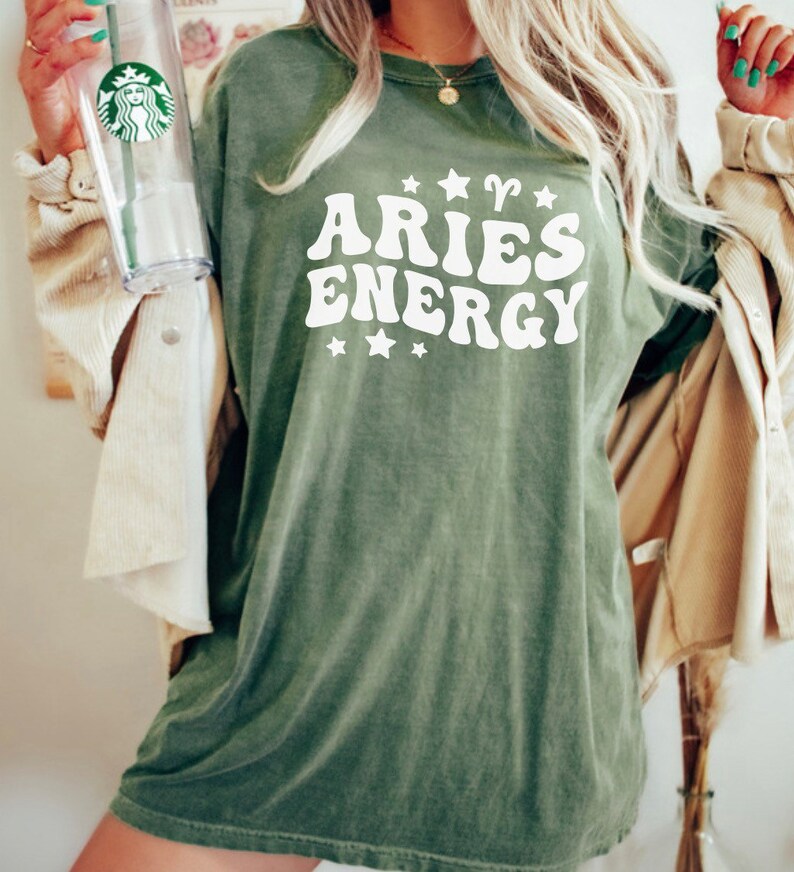 May include: Olive green t-shirt with the text "ARIES ENERGY" in white, featuring stars and the Aries symbol. The shirt has a vintage, slightly faded look. A clear Starbucks tumbler is held in the left hand.