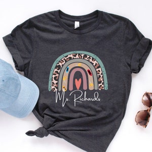 May include: Dark gray t-shirt featuring a rainbow design with scissors, a sewing machine, and a heart. The rainbow has a leopard print pattern. The text "Ms. Richards" is printed below the rainbow. A light blue cap and sunglasses are in the image.