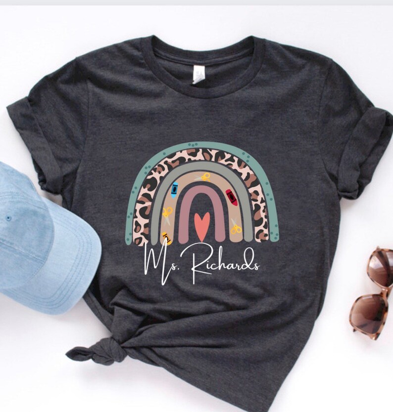 May include: Dark gray t-shirt featuring a rainbow design with scissors, a sewing machine, and a heart. The rainbow has a leopard print pattern. The text "Ms. Richards" is printed below the rainbow. A light blue cap and sunglasses are in the image.