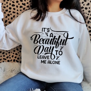 May include: White sweatshirt with the phrase "It's a Beautiful Day to Leave Me Alone" in black script. The sweatshirt is a crew neck style and is made of a soft material. The text is centered on the front of the sweatshirt.