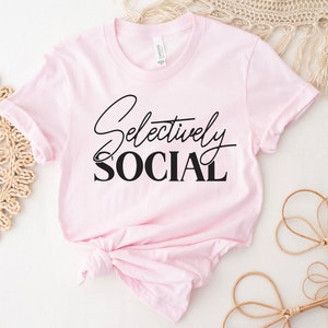 May include: A pink t-shirt with the text "Selectively Social" in black lettering.