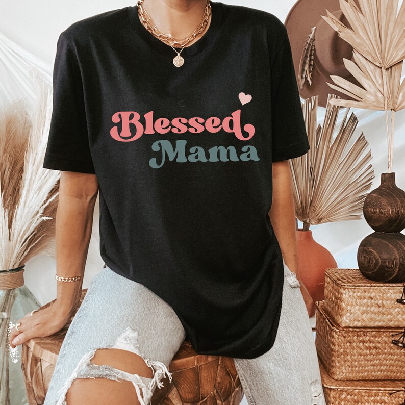 Puede incluir: Una camiseta negra con un gr&aacute;fico de estilo retro que dice "Blessed Mama" en letras rosa y azul con un peque&ntilde;o coraz&oacute;n rosa sobre el texto.