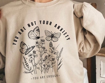 Può includere: Felpa beige con testo nero che recita "You are not your anxiety - You are enough" con un design floreale e farfalle.