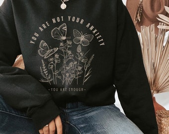 Può includere: Felpa nera con grafica beige di farfalle e fiori. Il testo recita "YOU ARE NOT YOUR ANXIETY" sopra il disegno e "- YOU ARE ENOUGH -" sotto. Fa parte di una collezione di abbigliamento.