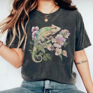 May include: A dark grey t-shirt featuring a graphic print of a green chameleon perched on pink and purple flowers. The chameleon has a long tail and is surrounded by green leaves.