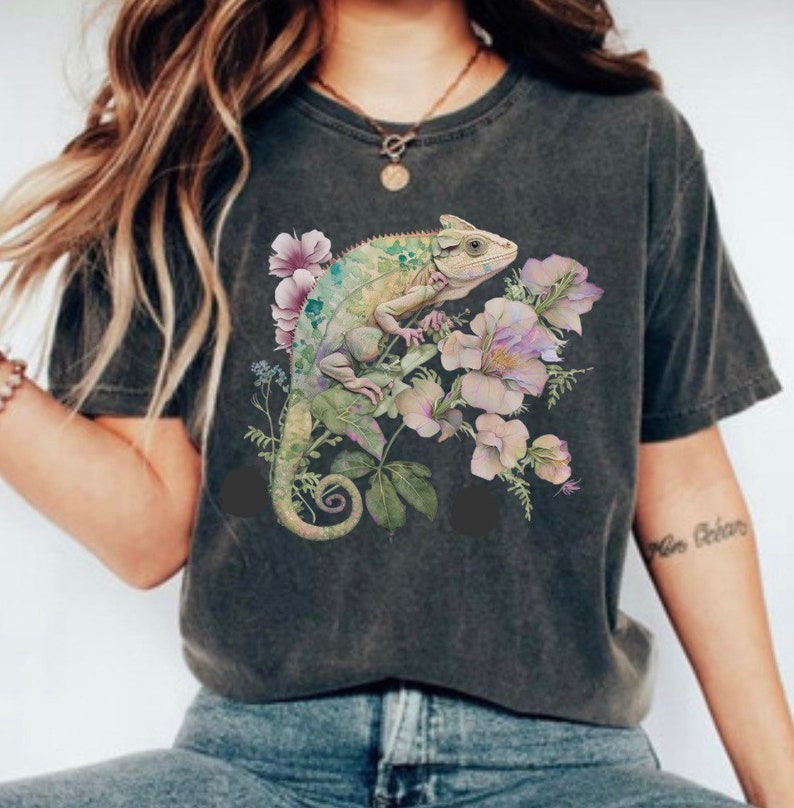 May include: A dark grey t-shirt featuring a graphic print of a green chameleon perched on pink and purple flowers. The chameleon has a long tail and is surrounded by green leaves.