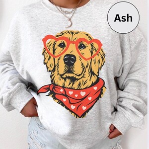 May include: Ash-colored sweatshirt featuring a golden retriever wearing heart-shaped glasses and a red bandana with white hearts. The dog's image is centered on the sweatshirt. The word "Ash" is in a white circle in the upper right corner.