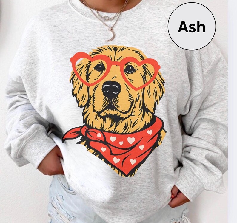 May include: Ash-colored sweatshirt featuring a golden retriever wearing heart-shaped glasses and a red bandana with white hearts. The dog's image is centered on the sweatshirt. The word "Ash" is in a white circle in the upper right corner.