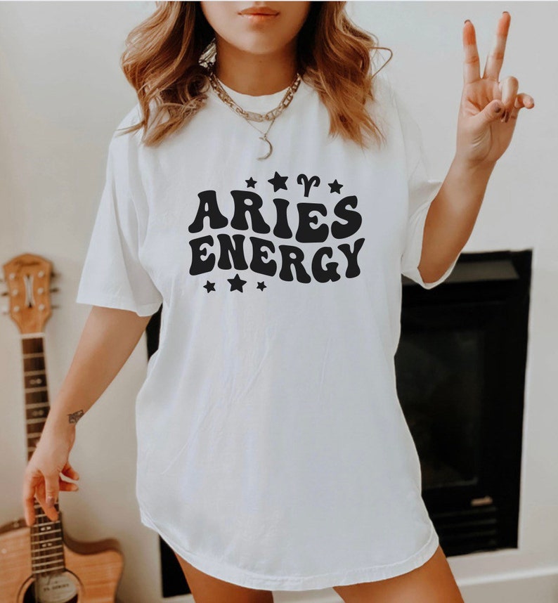May include: White t-shirt with the text "ARIES ENERGY" in a black, retro font, accompanied by star and Aries symbol graphics. The shirt is a casual, oversized fit.
