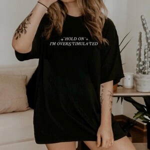 Może przedstawiać: Czarny, oversize'owy T-shirt z napisem "Hold On I'm Overstimulated" w kolorze białym.