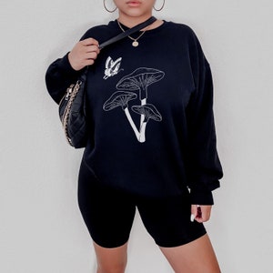 May include: Black sweatshirt with a white mushroom graphic and a butterfly design. The sweatshirt is paired with black biker shorts.