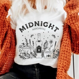May include: White t-shirt with black text that reads "MIDNIGHT MARGARITAS". The shirt features a black and white illustration of a bottle of tequila, a margarita glass, a mortar and pestle, and various herbs and flowers.