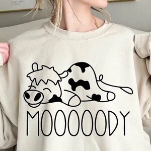 Peut inclure: Sweat-shirt beige avec un dessin de vache de dessin anim&eacute; noir et blanc. La vache est allong&eacute;e avec un visage souriant et des taches noires. Le mot "MOOOOODY" est imprim&eacute; en lettres noires en dessous.
