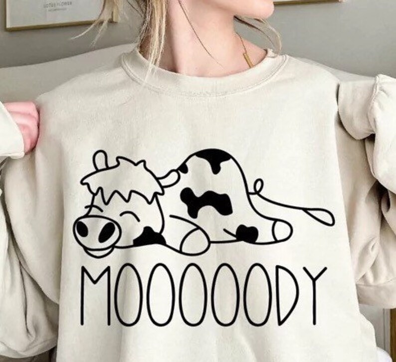Peut inclure: Sweat-shirt beige avec un dessin de vache de dessin anim&eacute; noir et blanc. La vache est allong&eacute;e avec un visage souriant et des taches noires. Le mot "MOOOOODY" est imprim&eacute; en lettres noires en dessous.