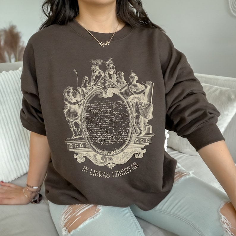 May include: A dark brown sweatshirt with a cream-coloured ornate design. The design features figures, script, and the words "IN LIBRAS LIBERTAS". The sweatshirt has a crew neck and long sleeves. The design is centered on the front.