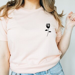 K&ouml;nnte beinhalten: Ein hellrosa T-Shirt mit einer schwarzen Silhouette einer Rose auf der linken Brust.