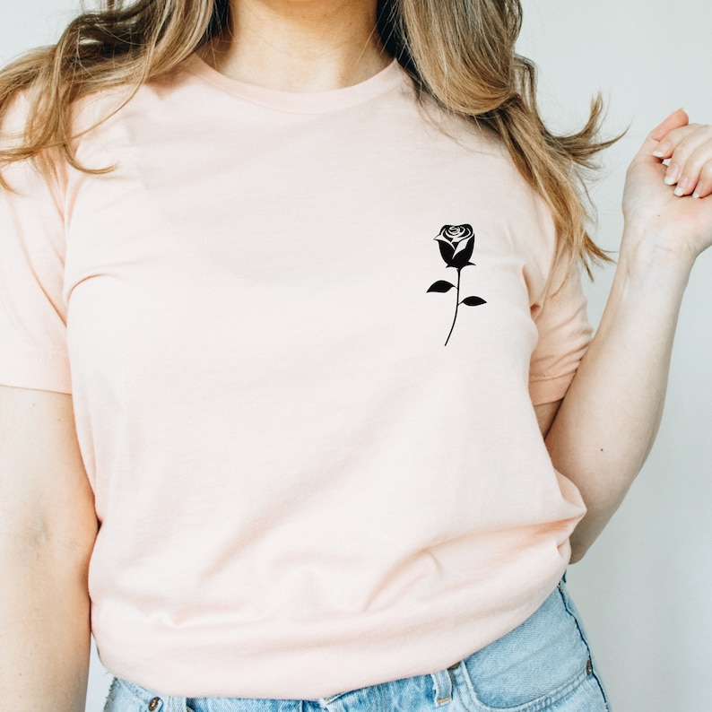 K&ouml;nnte beinhalten: Ein hellrosa T-Shirt mit einer schwarzen Silhouette einer Rose auf der linken Brust.