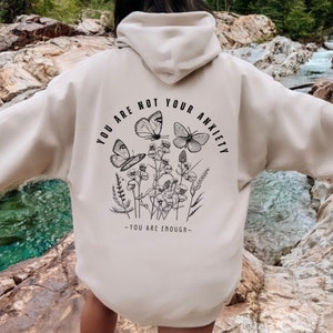 May include: A cream-colored hoodie with a black graphic print of butterflies and flowers. The text "You are not your anxiety - You are enough -" is printed on the back of the hoodie.