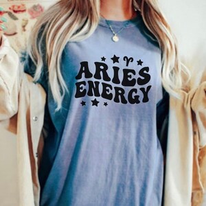 May include: A light blue t-shirt with the words "ARIES ENERGY" in black, stylized lettering. The design includes star and Aries symbol graphics. The shirt is a casual style.