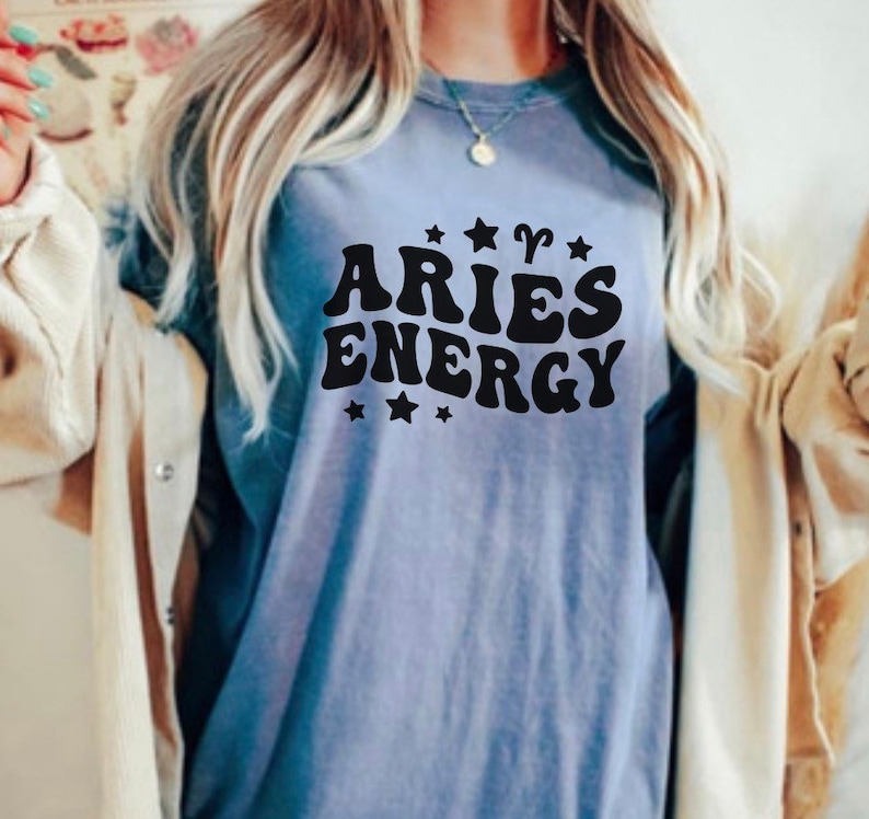 May include: A light blue t-shirt with the words "ARIES ENERGY" in black, stylized lettering. The design includes star and Aries symbol graphics. The shirt is a casual style.