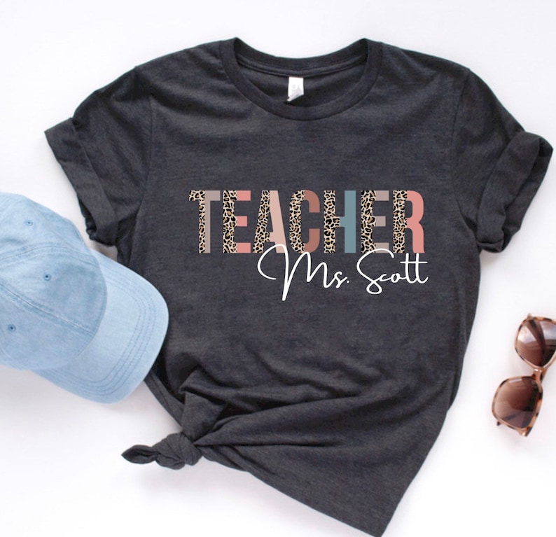 Pode incluir: Uma camiseta cinza escura com a palavra "TEACHER" em letras coloridas com um padr&atilde;o de leopardo. O texto "Mrs. Scott" est&aacute; escrito em branco abaixo da palavra "TEACHER".