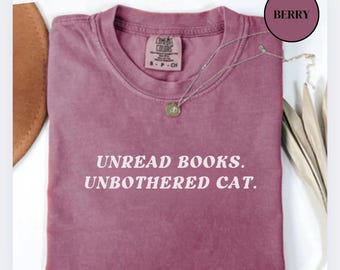 Może przedstawiać: Koszulka w kolorze jagodowym z nadrukiem "Unread Books. Unbothered Cat." w kolorze białym.