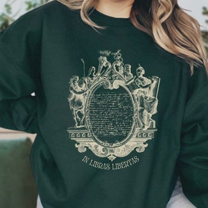 May include: A dark green sweatshirt featuring a vintage-style graphic print of a crest with figures and text. The text reads "In Libras Libertas".