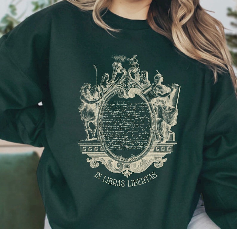 May include: A dark green sweatshirt featuring a vintage-style graphic print of a crest with figures and text. The text reads "In Libras Libertas".