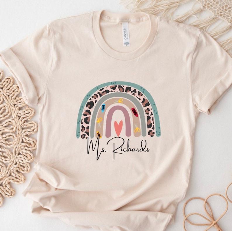 May include: Cream-colored t-shirt featuring a rainbow design with school supplies and a heart. The rainbow includes a leopard print section. The text "Ms. Richards" is written below the rainbow in a cursive font.