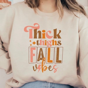 May include: A cream-colored sweatshirt with the text "Thick thighs fall vibes" printed in brown, orange, and pink.