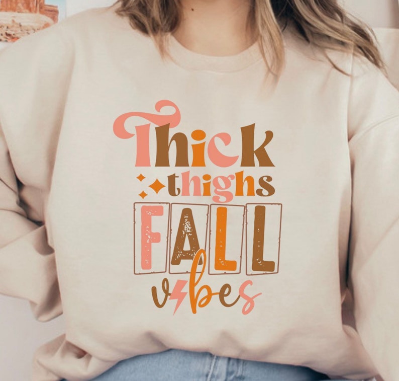 May include: A cream-colored sweatshirt with the text "Thick thighs fall vibes" printed in brown, orange, and pink.