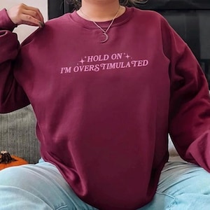 Może przedstawiać: Bordowa bluza z napisem "HOLD ON I'M OVERSTIMULATED" w r&oacute;żowych literach.