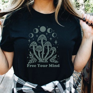 以下が含まれることがあります： 月の下にキノコが描かれた黒いTシャツ。Tシャツの下には「Free Your Mind」という文字が書かれています。