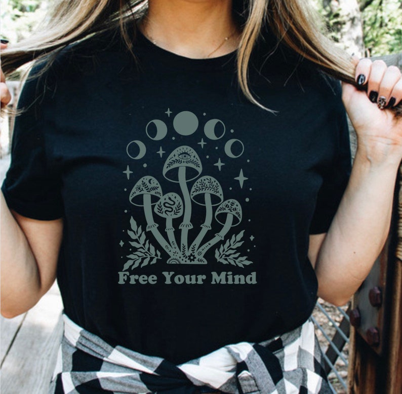 以下が含まれることがあります： 月の下にキノコが描かれた黒いTシャツ。Tシャツの下には「Free Your Mind」という文字が書かれています。