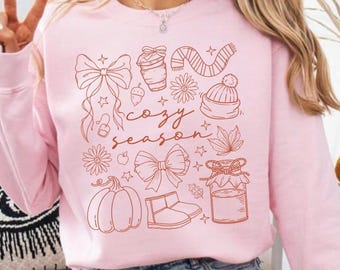 Könnte beinhalten: Ein hellrosa Sweatshirt mit einem braunen Grafikdesign mit herbstlichen Illustrationen, darunter ein Kürbis, eine Schleife und die Worte "cozy season."