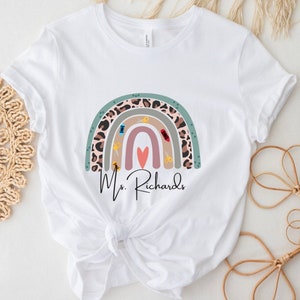 May include: White t-shirt featuring a colorful rainbow design with sewing-themed elements. The rainbow includes a leopard print section, and the name "Ms. Richards" is printed below the rainbow in a script font.