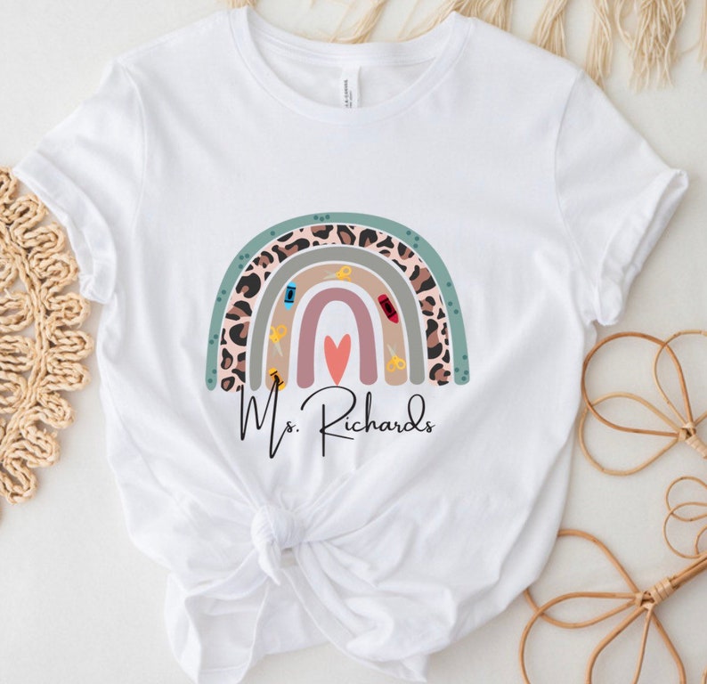May include: White t-shirt featuring a colorful rainbow design with sewing-themed elements. The rainbow includes a leopard print section, and the name "Ms. Richards" is printed below the rainbow in a script font.