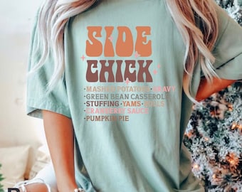 May include: A light blue t-shirt with the text "SIDE CHICK" in a retro font. The text is in shades of orange and brown. Below the text is a list of Thanksgiving side dishes: mashed potatoes, green bean casserole, stuffing, yams, rolls, cranberry sauce, and pumpkin pie.