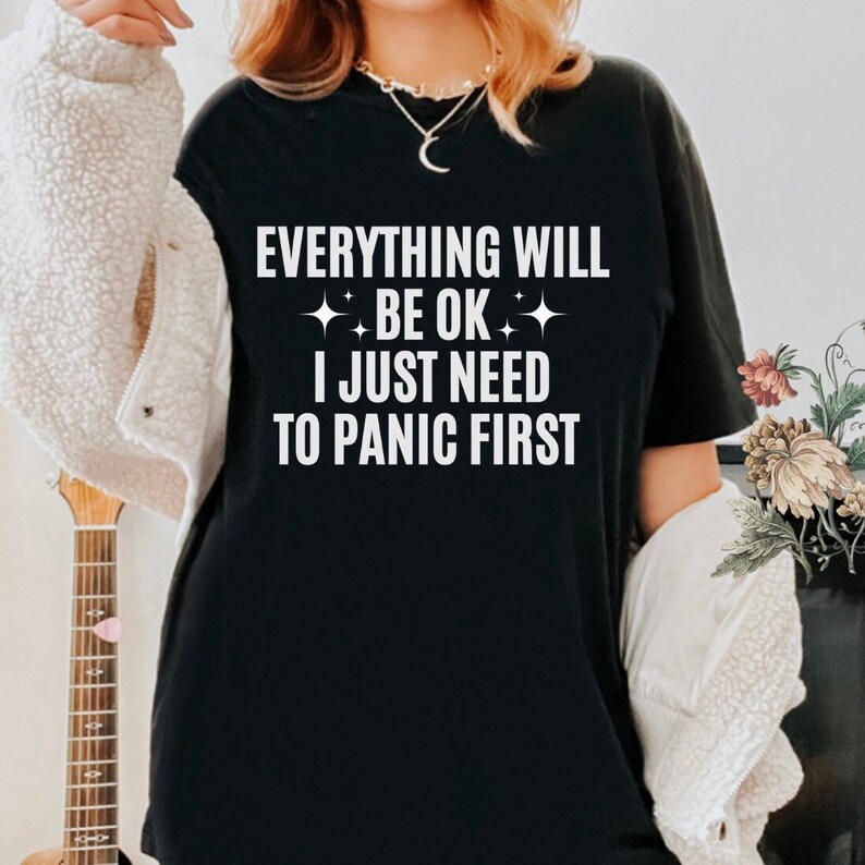 May include: Black t-shirt with the white text "EVERYTHING WILL BE OK I JUST NEED TO PANIC FIRST". The shirt has a crew neck and short sleeves. The text is in a bold, sans-serif font.